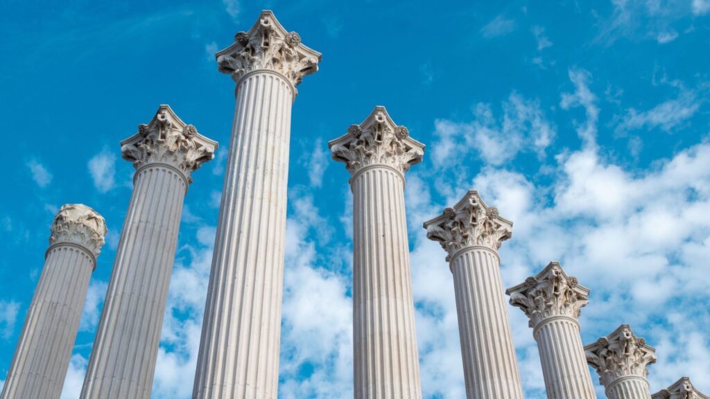 A low-angle view of a row of tall, white Corinthian stone columns reaching up toward a bright blue sky with scattered clouds.