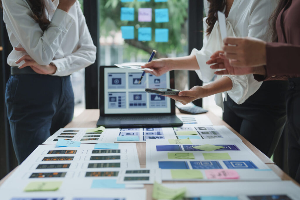 A design team standing around a table reviewing printed mobile app UX wireframes and sticky notes, with a laptop displaying similar layouts in the background.
