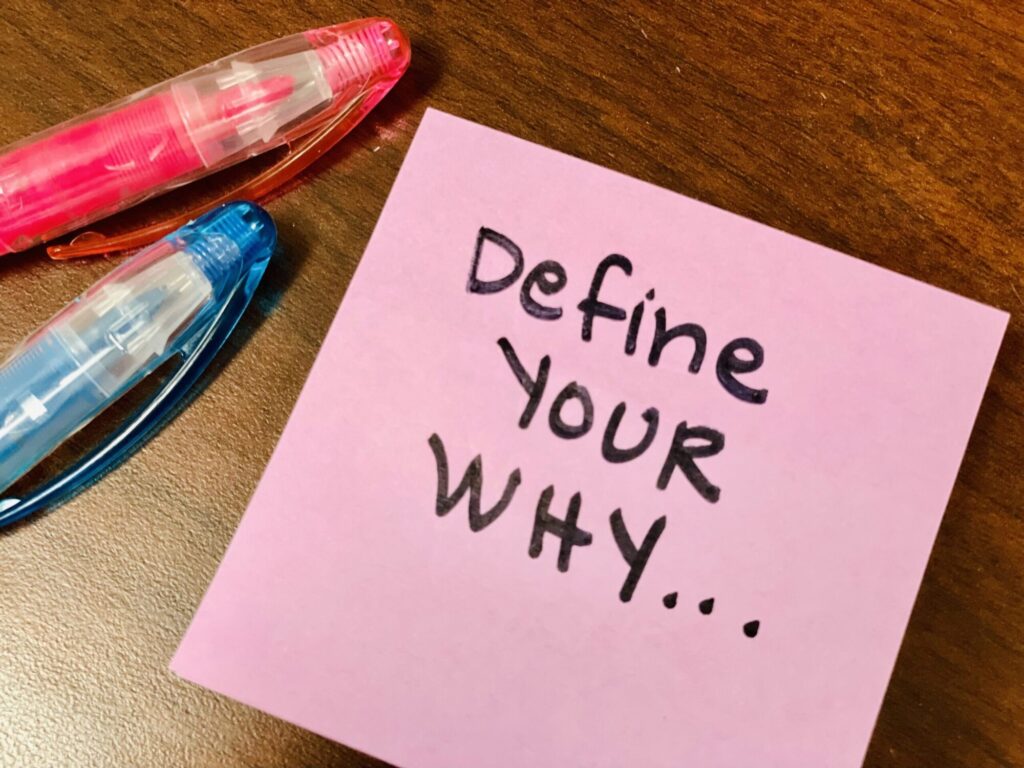 A pink sticky note on a wooden desk with the handwritten text "Define your WHY..." next to two pens.