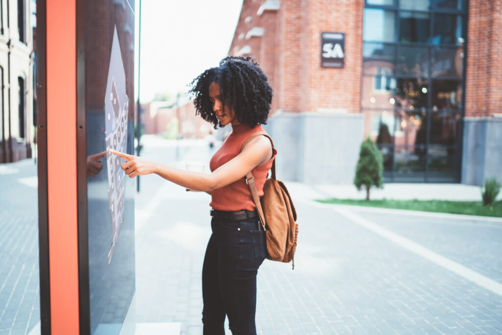 A woman with a backpack pointing at a digital interactive map kiosk on a paved city street.